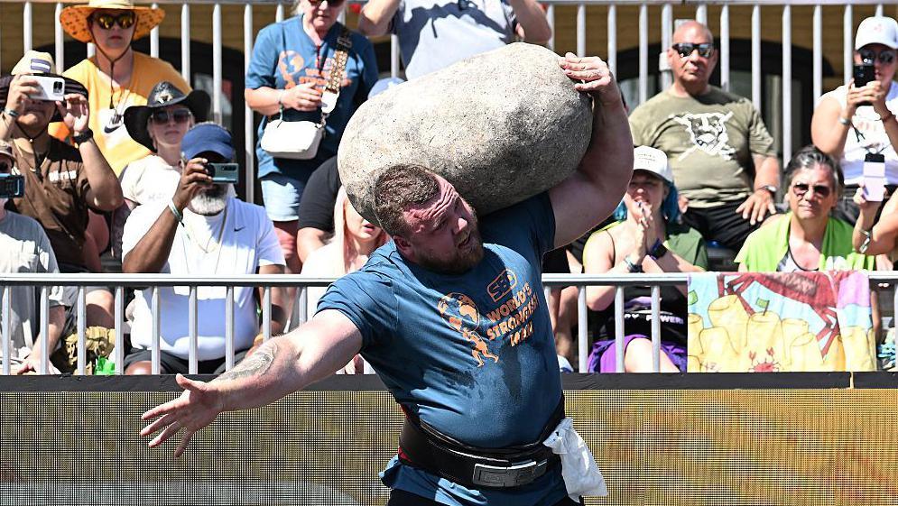 Tom Stoltman competing in World's Strongest Man. She is holding a large stone on his shoulder.