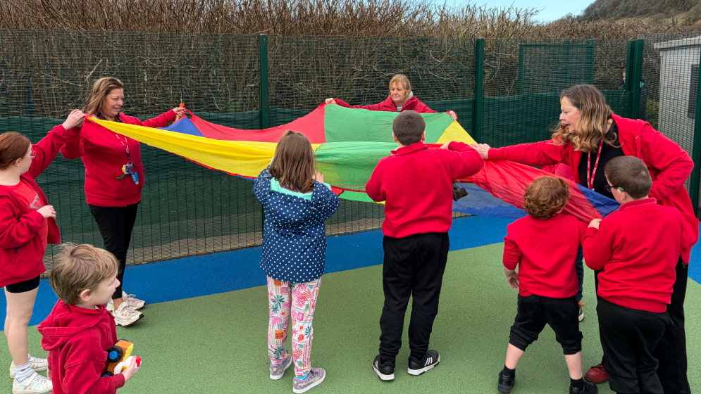 Grwp o blant mewn siwmperi coch ar iard ysgol yn dal canopi parachute lliwgar gydag aelodau staff yn helpu