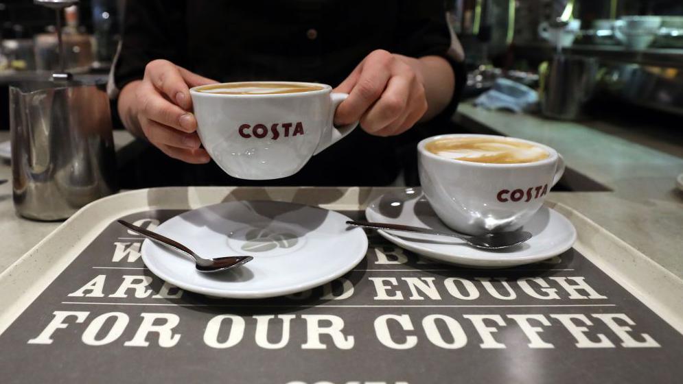 A barista putting two white Costa Coffee cups filled with coffee on to white coasters on a Costa branded tray
