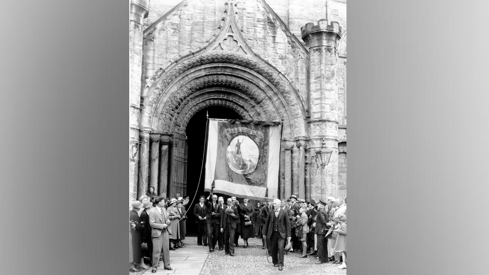 A black-and-white picture of people carrying a large banner out of a cathedral. They are dressed smartly in suits and dresses.