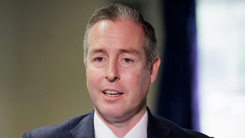 Paul Givan has salt and pepper coloured hair parted to the side and is clean shaven. He is wearing a dark grey suit jacket, white shirt and burgundy tie.