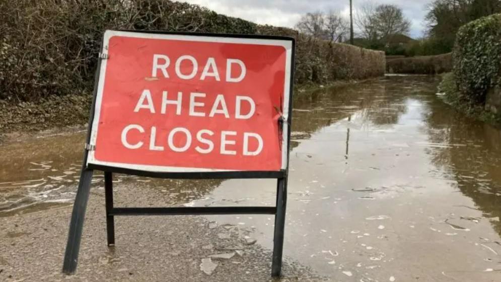 A red sign reads road ahead closed on a country, flooded road