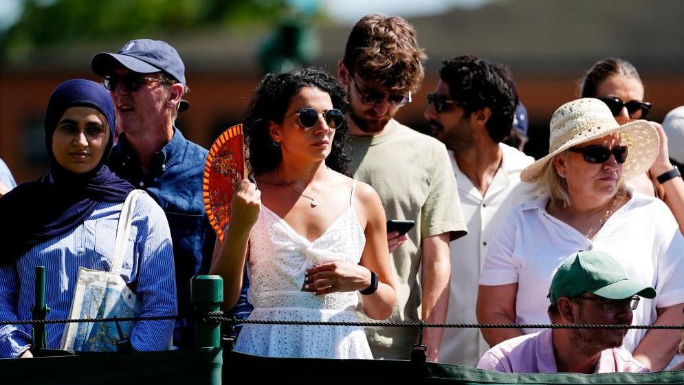 Several people standing in a crowd at Wimbledon in the heat. They are dressed in summer clothes, often in caps and sunhats. The woman in the centre of the picture is using a fan and wearing sunglasses.
