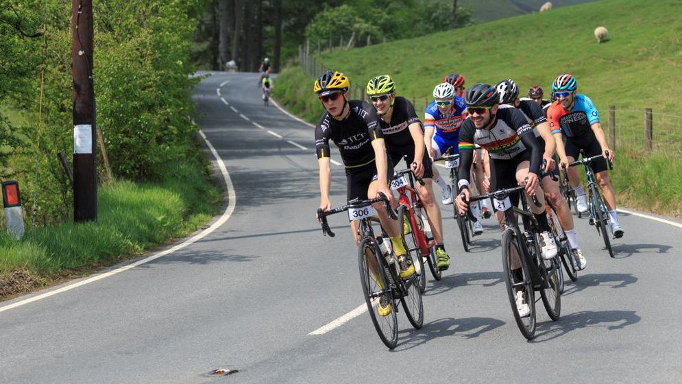There are several cyclists on the road. To the right and left there is a grassy verge, the cyclists are on the right-hand side of the road.