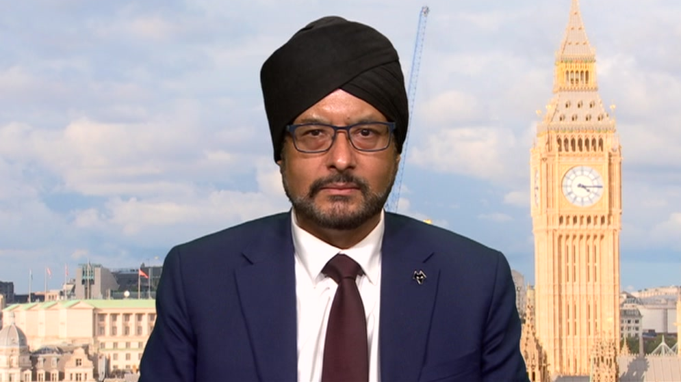 Mr Juss is wearing a dark suit and turban and glasses. He is in a TV studio for the interview with an image of Big Ben behind him.