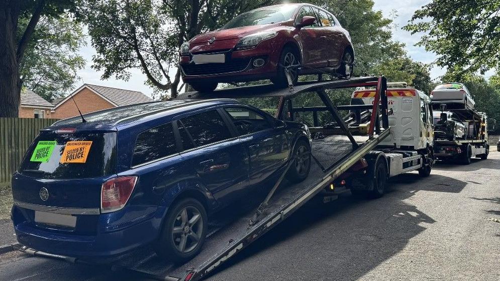Four vehicles, on two transporters, on a road, with trees on it. There is a blue and a red vehicle on the back of the white lorry.