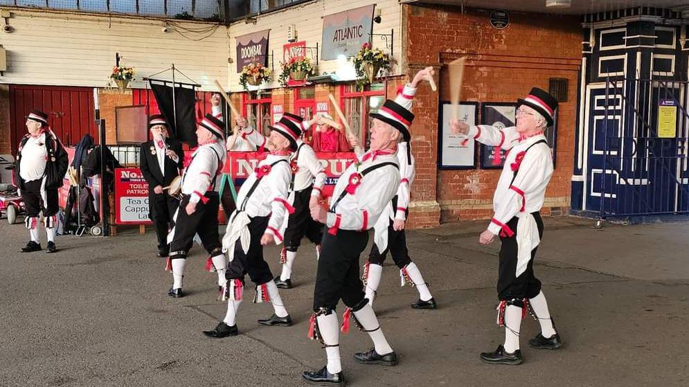 A group perform a morris dance on the platform at Cleethorpes station. The dancers are wearing bells around their ankles and are waving wooden sticks.