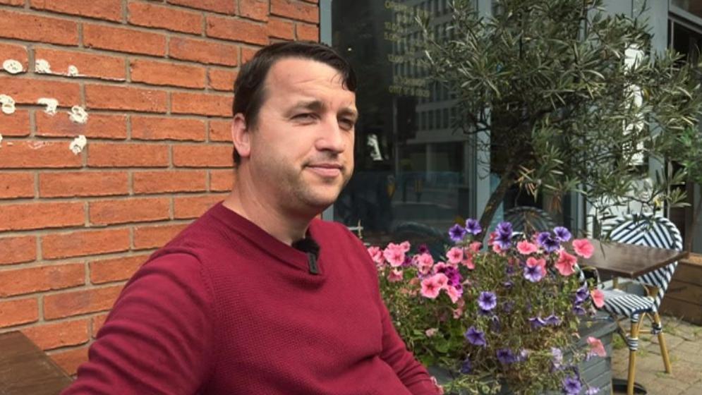 A man wearing a burgundy jumper in front of a brick wall and plants