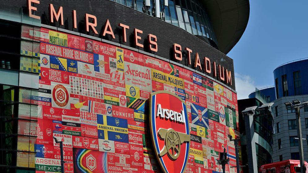 An exterior view of Arsenal's Emirates Stadium, showing a giant club badge surrounded by flags and banners from supporter groups from across the world