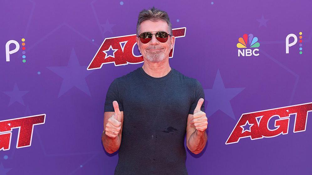 Simon Cowell smiling and giving a thumbs up with both hands. He is wearing sunglasses and a black t-shirt.