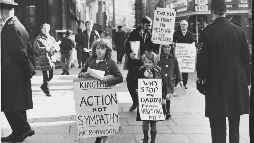 A black and white photo of a demonstration. Children are holding signs reading: "King Hill action not sympathy Mr Robinson", "Why stop my Daddy from visiting me?" and "Two men in court for helping the homeless!". Behind them a man's sign reads: "Tory council + Labour government. Broken families at King Hill." A policeman is watching on. There is a double decker bus in the background. 