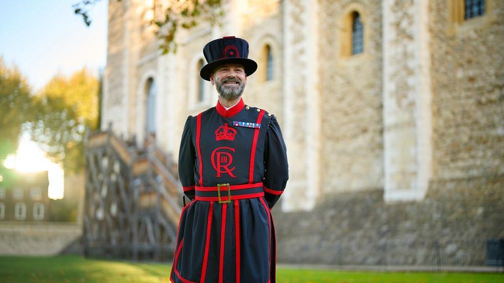 Ryan Brown with a short cropped beard smiling at the front of the tower of London. He is wearing a ceremonial blue and red tunic with a belt around its waist.  He is wearing a tall blue hat. The hat is trimmed with red. A Good Conduct medal for long service is pinned on the chest of his tunic which is a Yeoman Warder uniform. 
