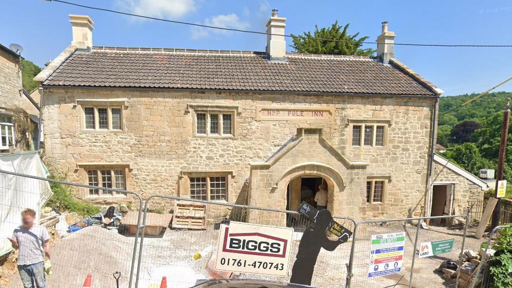 The Hop Pole Inn in Limpley Stoke on a sunny day while construction work is being carried out on it. It is an old two storey brick building with a pointed archway entrance and small, sunken sash windows. Outside the front there is metal heras fencing blocking the pub off from the road, a small skip, wooden pallets, and a huge pile of stones. 