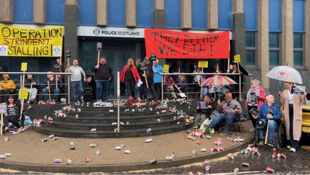 A group of campaigners with signs saying Operation Stalling and They Dither, We Die outside a police building. Cans are strewn across the ground, a reference to the allegation the police are kicking the can down the road