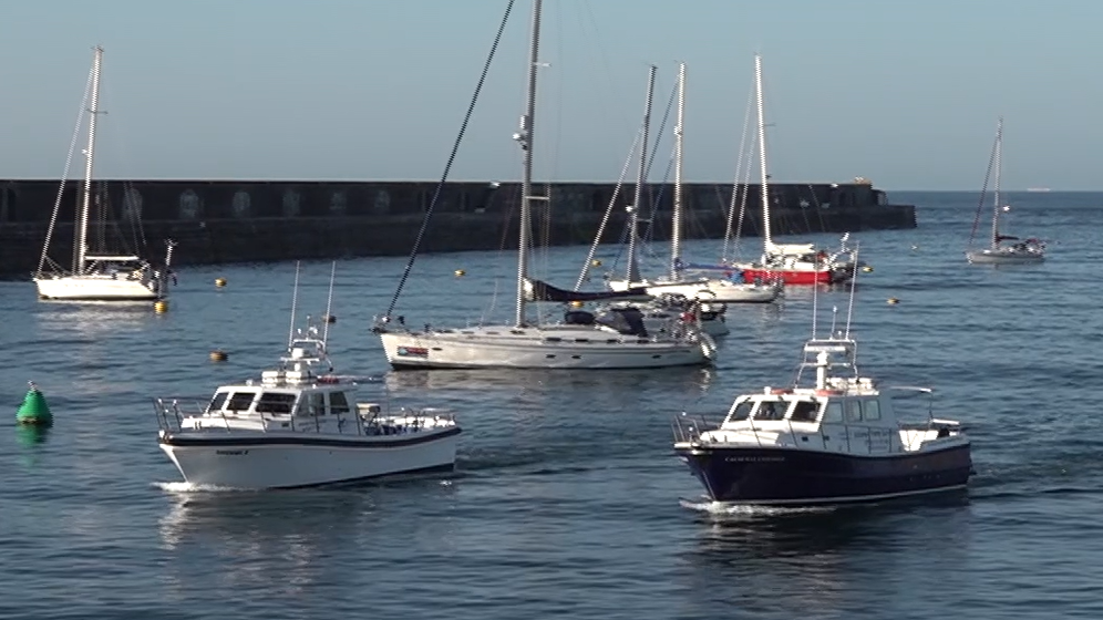 Sorcerer II and Causeway Explorer are sailing side by side into the harbour on a bright sunny day. There are five yacht moored inside the harbour wall.