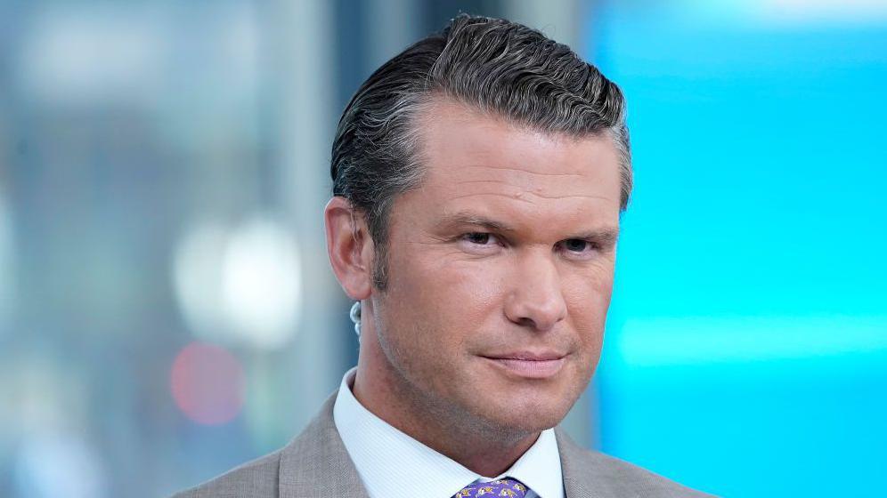 Pete Hegseth in a grey suit wearing an earpiece looks at the camera