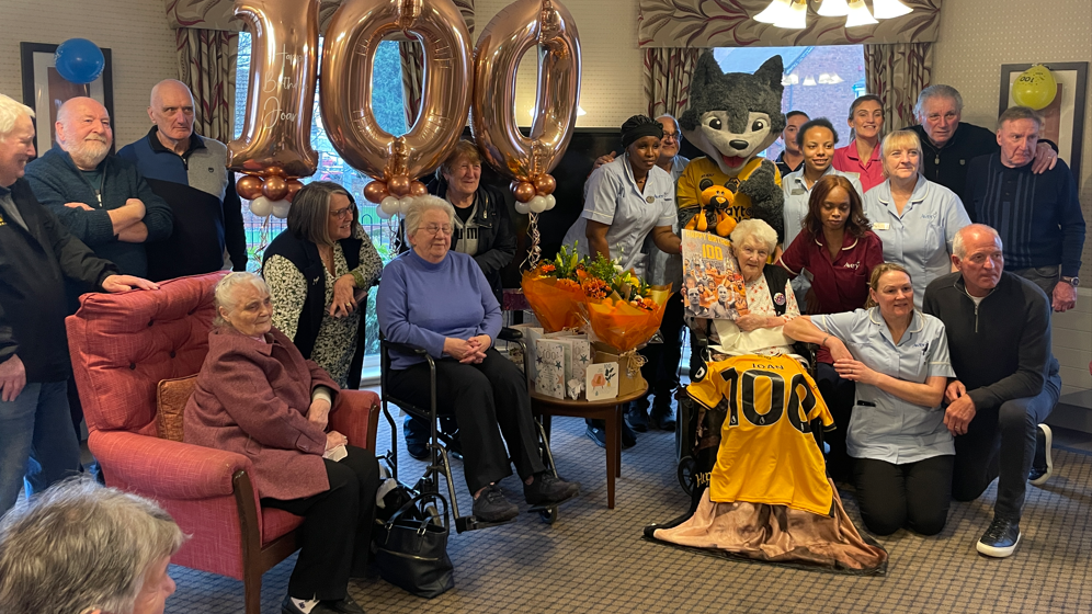 Joan is sat in a chair with a Wolves shirt draped over her lap and she is holding a birthday card. She is surrounded by former players, club associates, care home staff and friends.