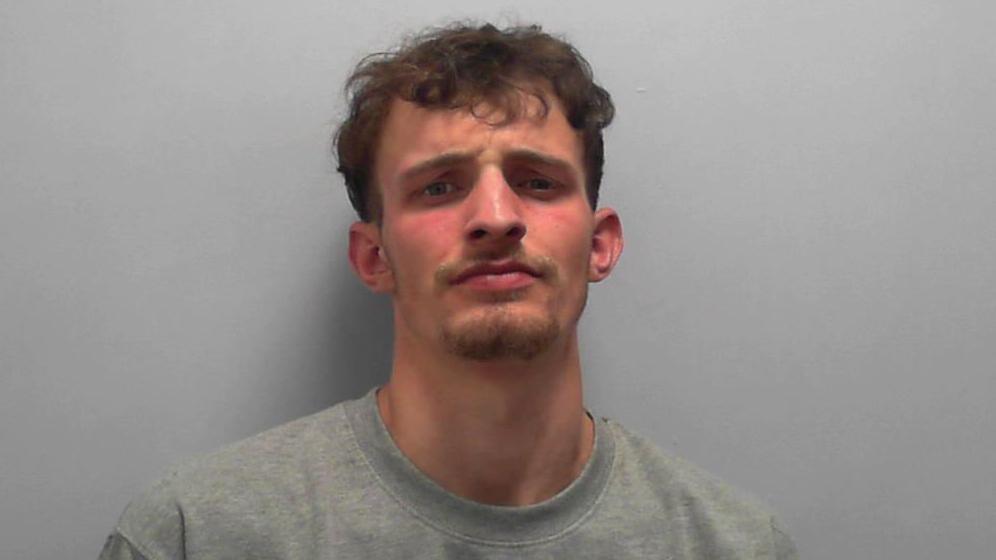 Frankie Lee Morrison's police mugshot - against a plain background, a young man stands wearing a grey jumper. He has short curly brown hair and a goatee beard.