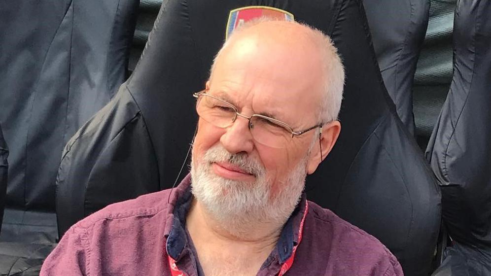 Jeffrey Holmes looks away from the camera as he sits in a large black chair. He is bald with some grey hairs on his head and he has a grey beard. He wears glasses along with a burgundy shirt with a lanyard around his neck.