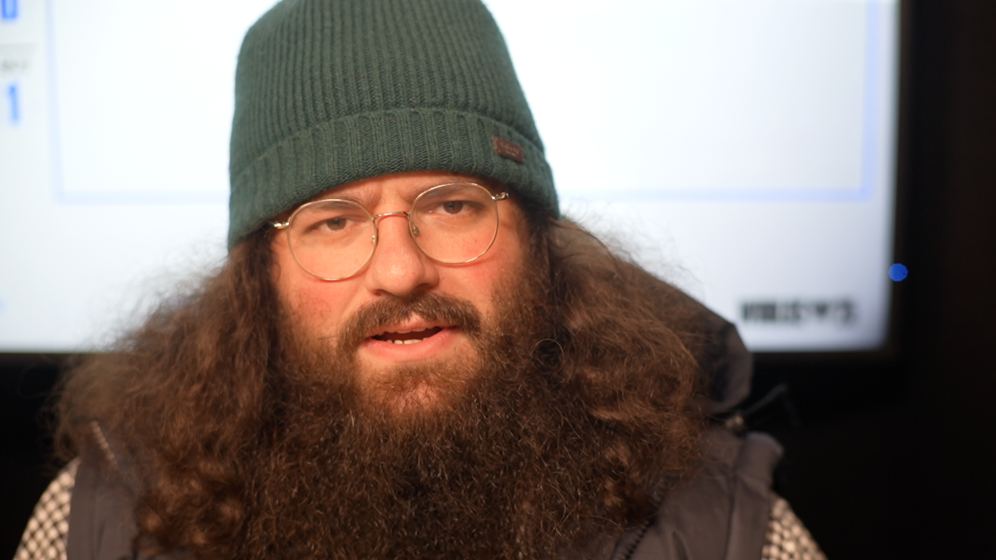 Edward Hale was wearing a green hat and looking at the camera. He had long ginger hair and a long beard, and was wearing glasses and a chequered shirt.