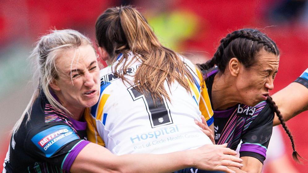 Leeds player Ruby Enright, with a white shirt bearing the number one, is tackled by St Helens players Jodie Cunningham (left) and Zoe Harris (right), both wearing black with purple trim,