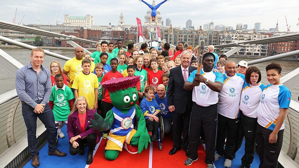 A large group of people, mainly children, wearing Glasgow 2014 t-shirts. They are gathered on a bridge. In the middle is Clyde the thistle.