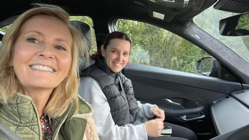 Two women at sitting in the front of a parked car. They are both looking at the camera and smiling. One woman is blonde and the other is a brunette. 