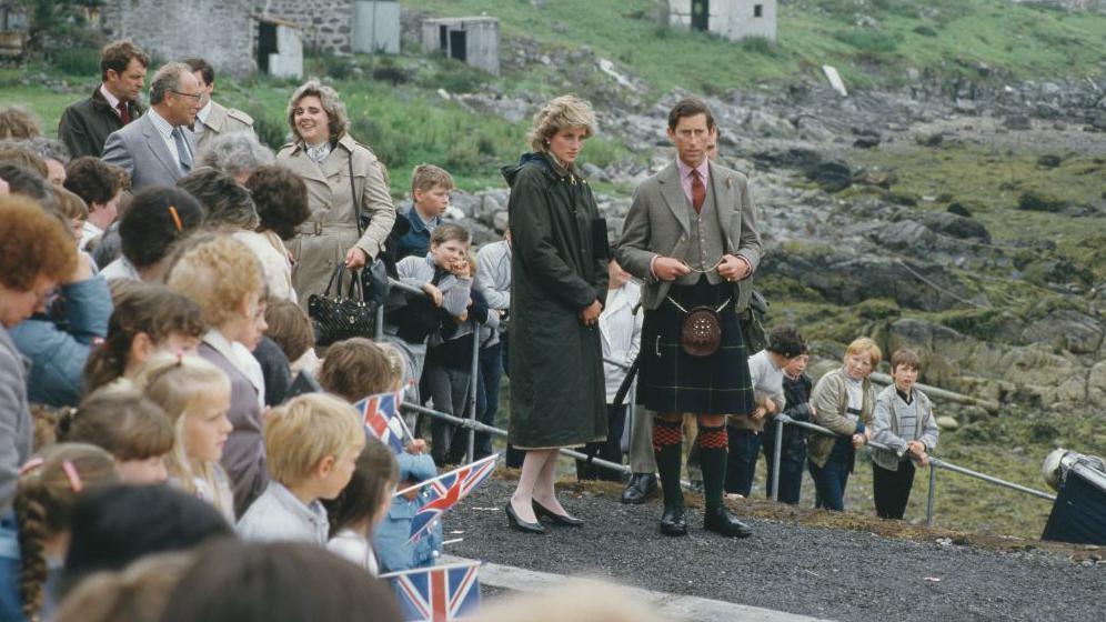 Diana is wearing a long, green raincoat. She is standing next to Charles who is wearing a kilt and a tweed jacket and waist coat. There islanders gathered around them.