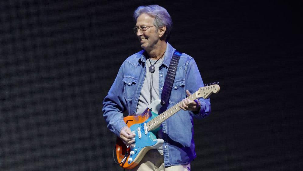 Eric Clapton with an orange and blue guitar performing on stage.