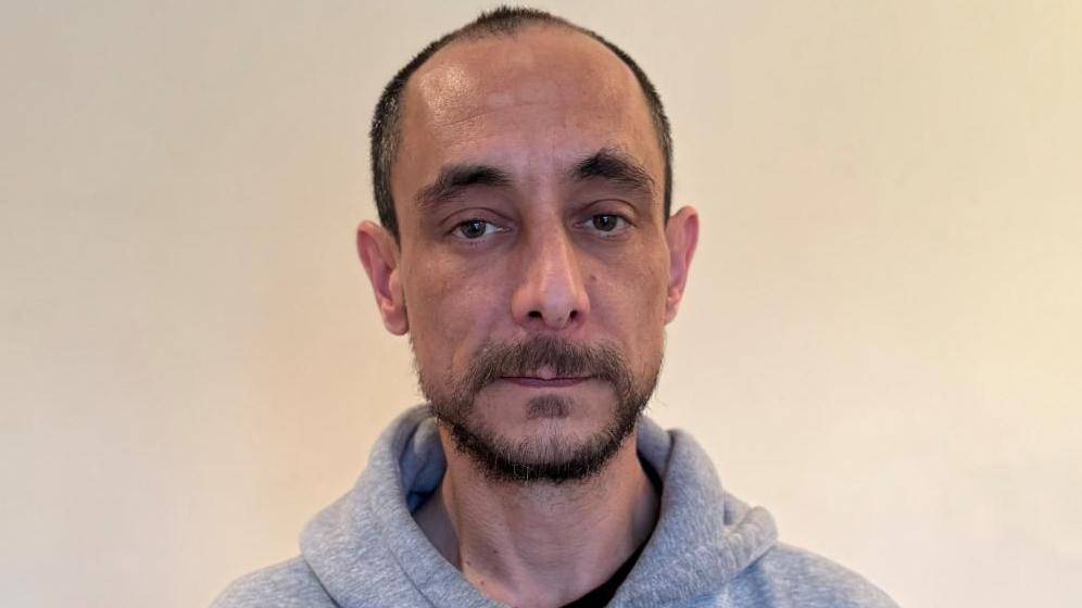 Steve looks at the camera and wears a grey hooded jumper. He has a dark beard, brown eyes and is partly bald. The background is blank.