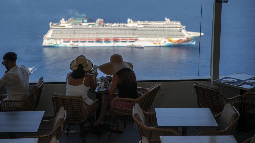 Two women wear sun hats, turned away from the camera looking over the Aegean sea as a cruise ship floats by.