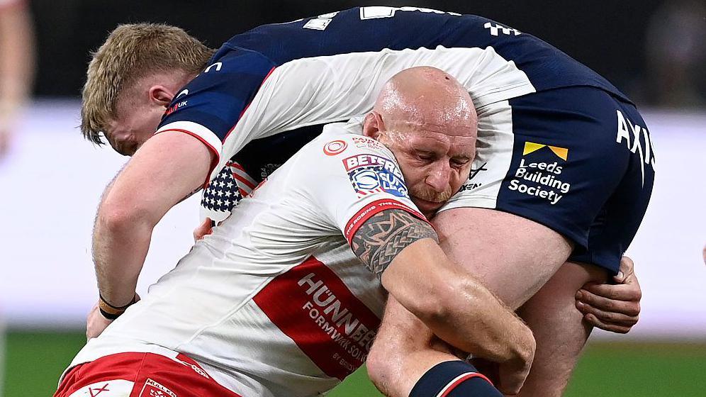 Hull Kingston Rovers forward Dean Hadley, in white and red, lifts Leeds Rhinos' James McDonnell, in dark blue and white, in a tackle.