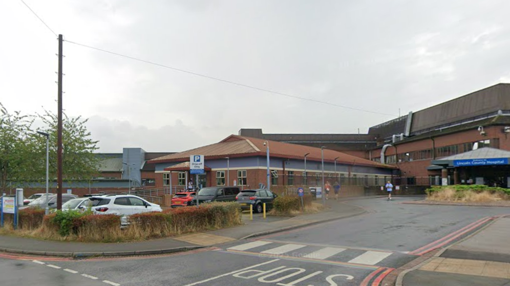 Exterior shot of the main entrance to Lincoln County Hospital. It is a red-bricked building with a blue sign over the main doors. There are other buildings in the background, as well as parked cars.
