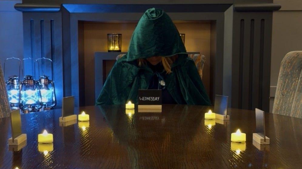 A woman in a green velvet hood is sat at a dining table with tealights and a card in front of her with the word Wednesday