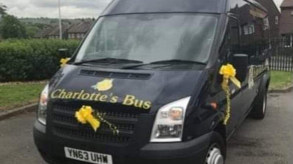 The black minibus has Charlotte's Bus painted on the front in yellow letters. Yellow ribbons are attached to the front and the wing mirrors. 