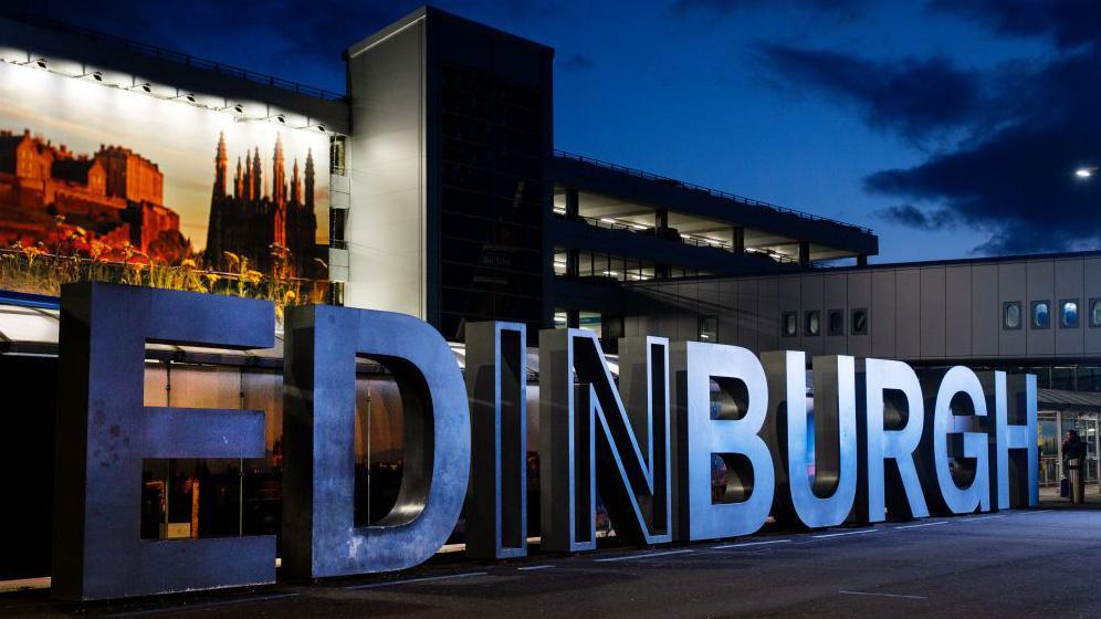 Large metal letters spell out "Edinburgh" in capitals outside the airport. It is dark and there are lights on in a multi-storey car park.