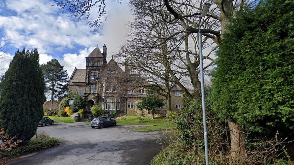 A Victorian-style with multiple storeys in the middle distance, surrounded by trees and behind a car park