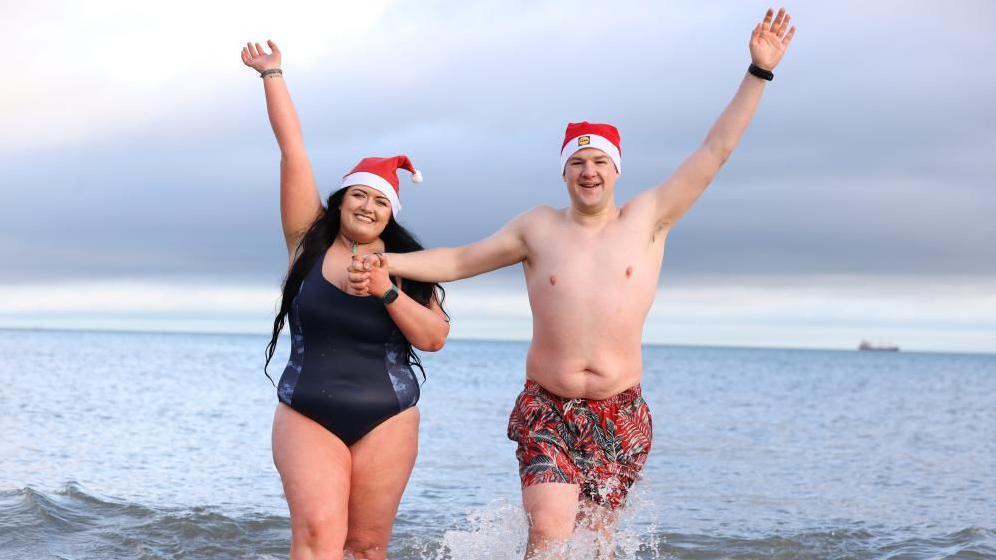 Rebecca, left, is wearing a navy swimming costume and red and white Santa hat, she has her arm up in the air. She is holding hands with Joel, right, who has patterned swimming shorts n and a red and white Santa hat.