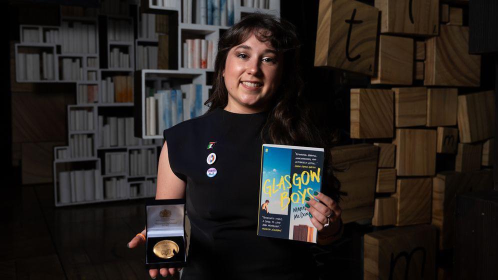 Margaret McDonald stands in a shadowy room with books on shelves in the background. She holds the Carnegie Medal and her book Glasgow Boys. She smiles at the camera and wears a black top with badges. She has long brown hair and a fringe.
