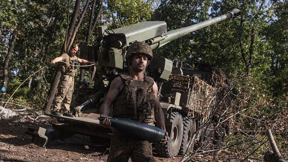 Two Ukrainian soldiers standing in front of a field gun. The one nearest to the camera is holding a shell.