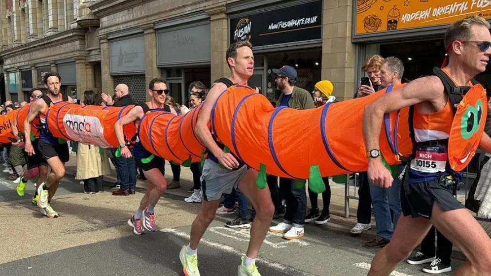 Men running in an orange caterpillar costume. They are joined at the chest and back by a concertina-type fabric.