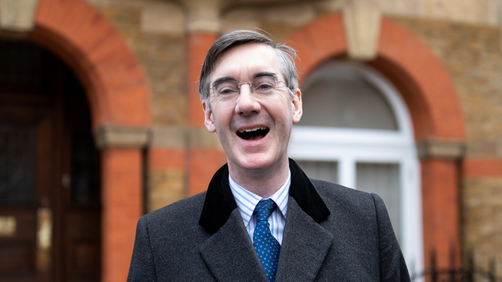 Jacob Rees-Mogg laughing. He is wearing a grey coat, blue tie and blue and white-striped shirt.