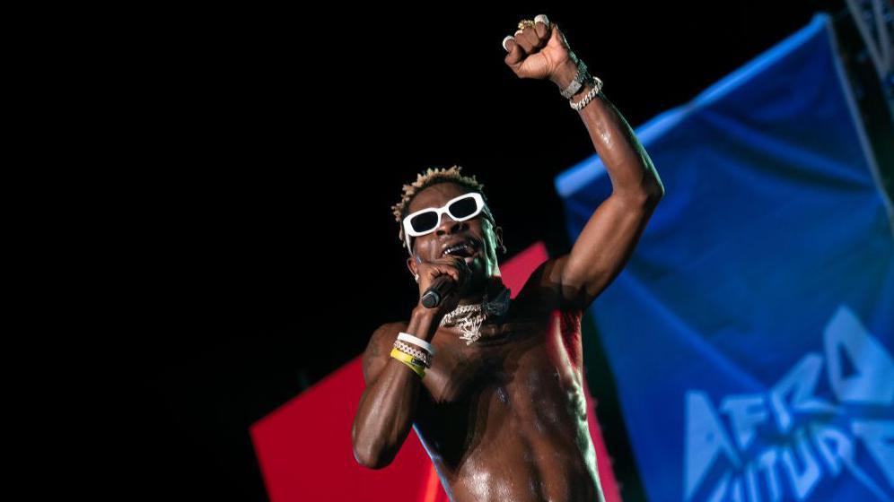 A bare-chested Shatta Wale singing at a concert. He is wearing white plastic sunglasses and has his fist raised.
