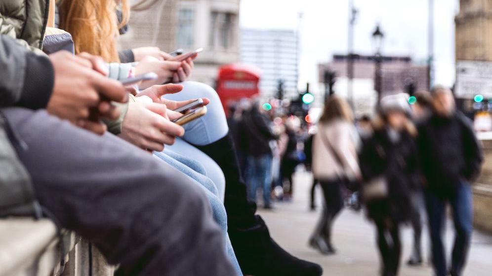 Close up of the hands of four young people holding their mobile phones while sitting, with a blurred image of people walking down the street and a red phone box in the background. 