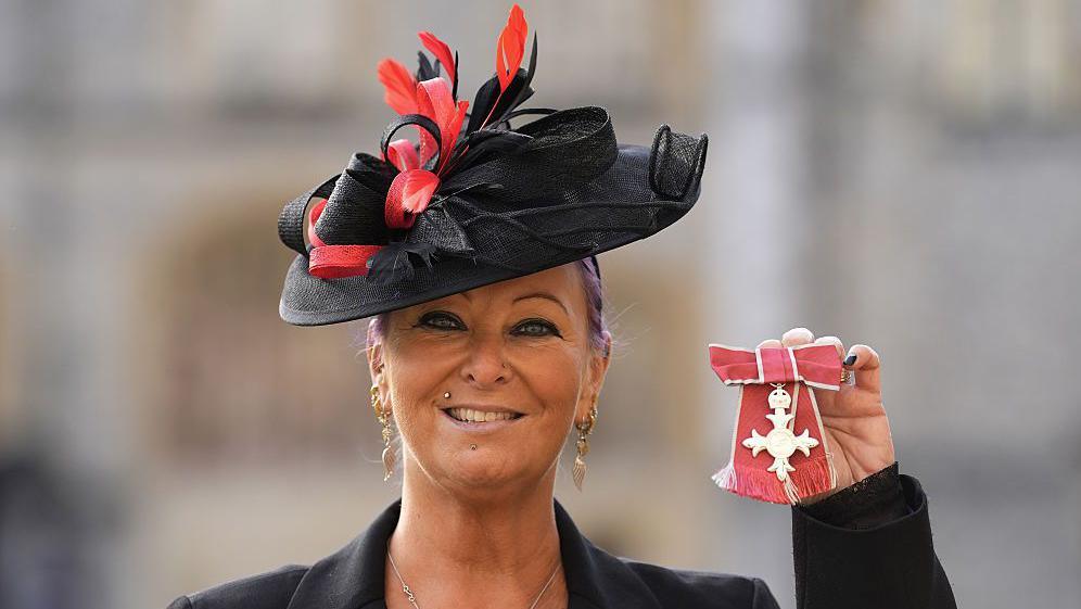 Charlotte Charles, wearing a black red hat, has purple tied-back hair and dangly earrings. She is holding an MBE medal in her right hand. She is wearing a black jacket and a necklace.