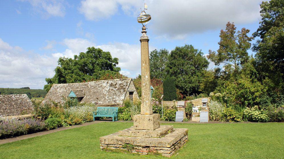 The sundial in The Armillary Court at Snowshill Manor in Gloucestershire in the centre of a square lawn of green grass with a bench in the background and a traditional cotswold building behind it. 