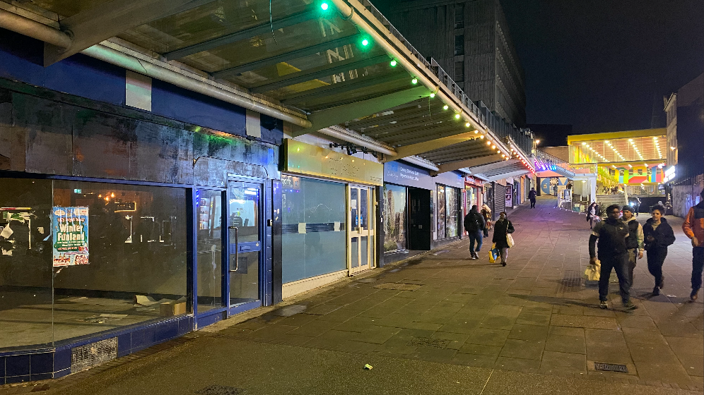 A view of a row of empty shops with people walking past in the dark and some lights in the distance