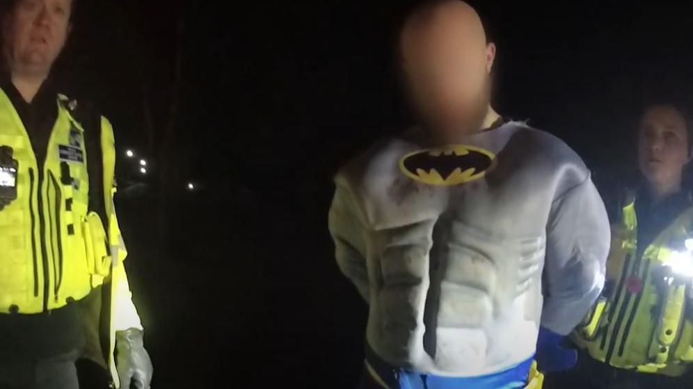 A man wearing a Batman costume with his hands behind his back stands next to two police officers