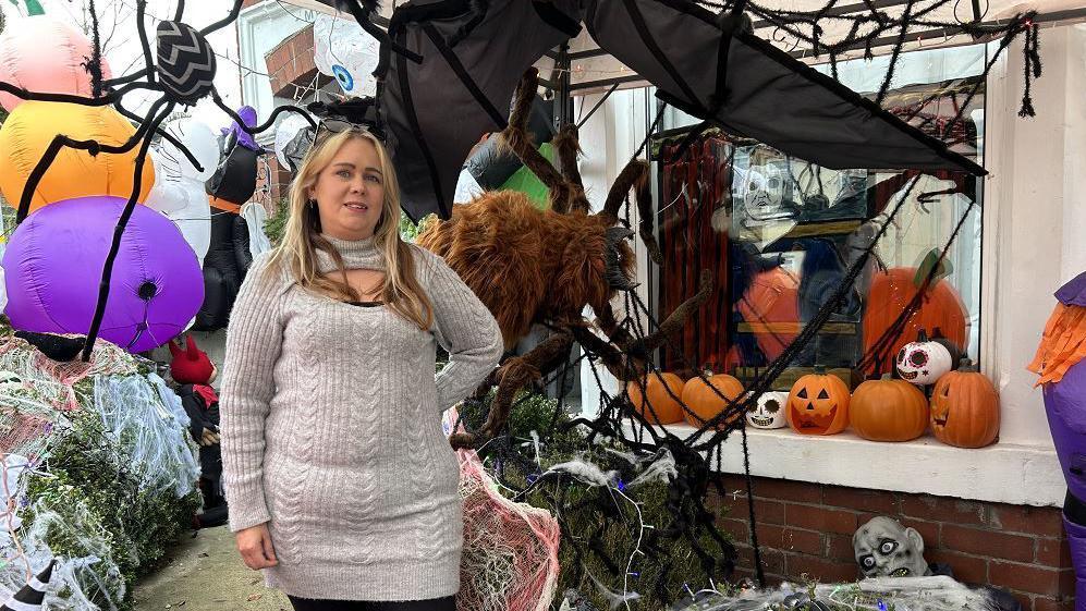 A woman standing in front of Halloween decorations on her house. They include inflatable pumpkins and scary masks and large fake spiders.