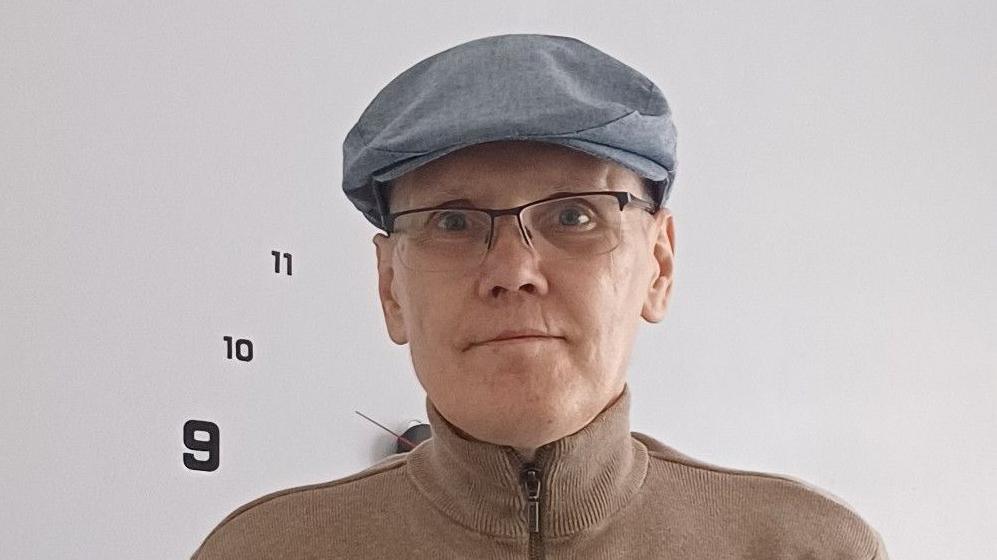 Robert Best is seen wearing glasses, a light blue flat cap, and a tan zip-up jumper stands in front of a white wall with a large minimalist clock showing the numbers 9, 10, and 11.